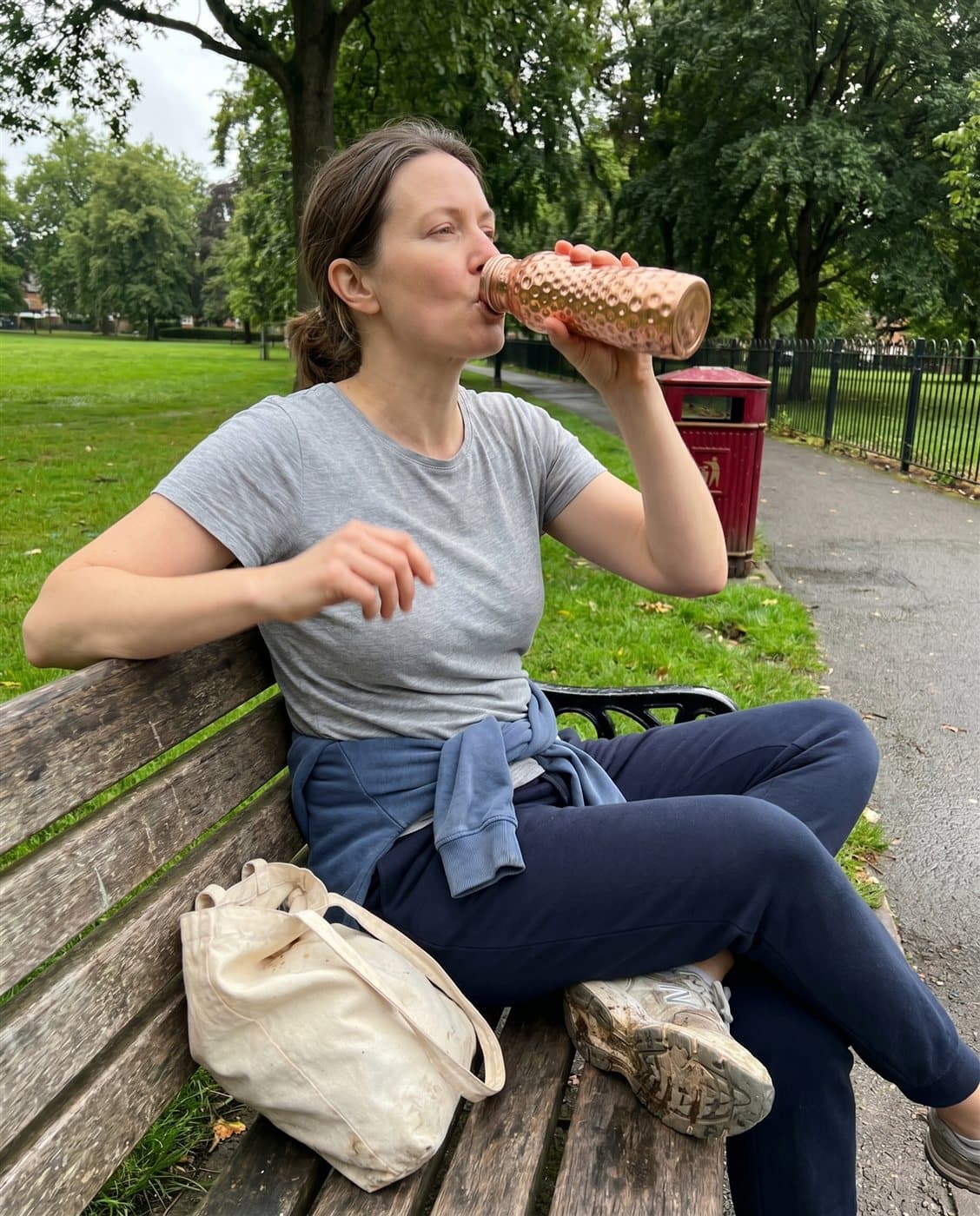 Someone taking a drink from the curved copper bottle on a park bench after a walk
